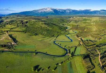 Aerial view of Livanjsko Polje with green meadows, a river and the mountains in the background.